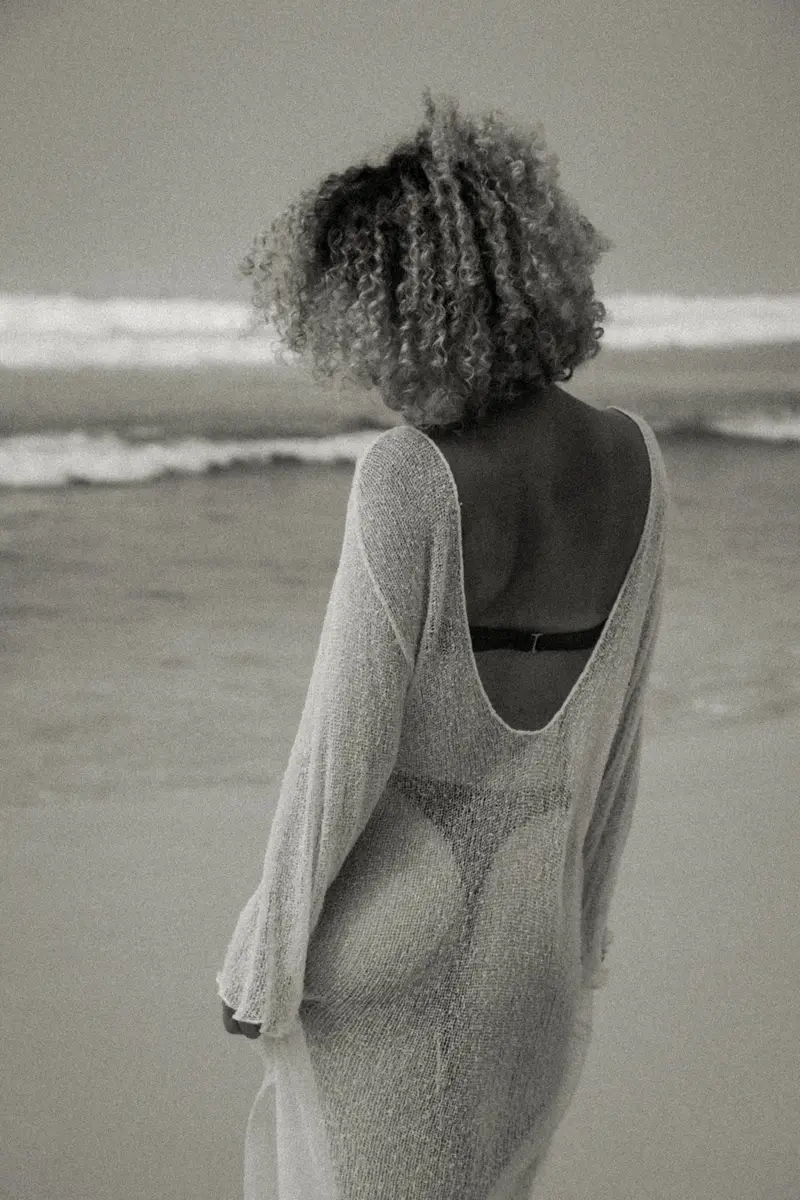 Femme aux cheveux bouclés, de dos, en robe au crochet face à la mer