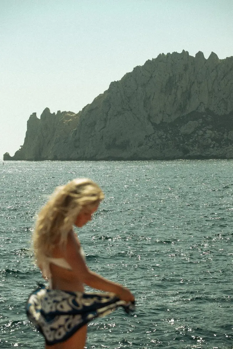 Femme blonde dans l'eau face à une falaise, un paréo à la main
